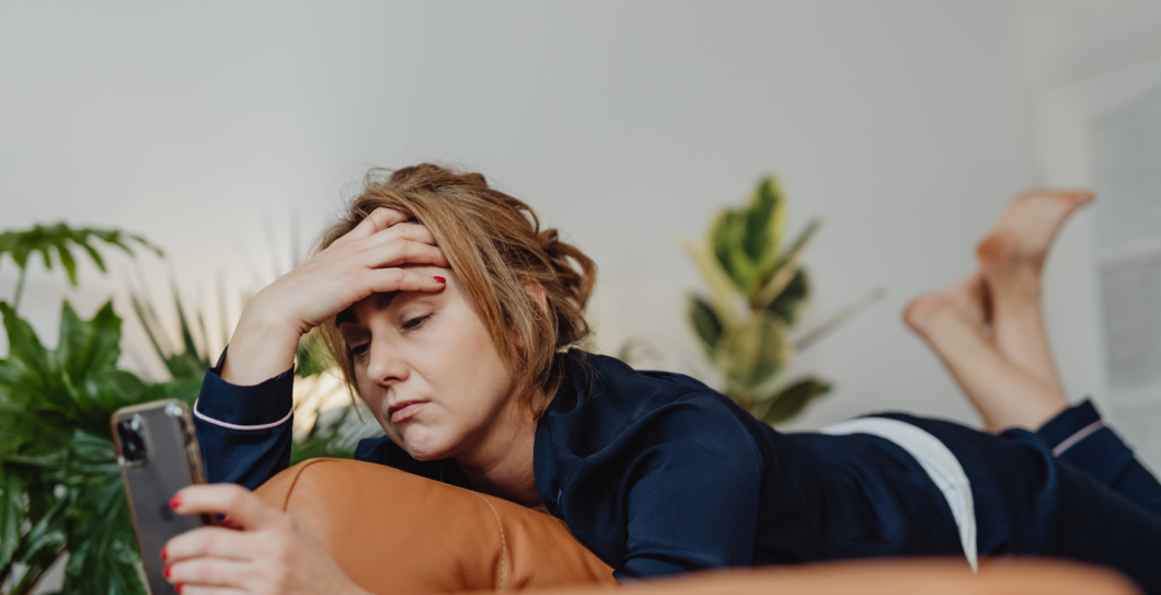 jonge vrouw ligt op de bank met mobiel in de hand en kijkt vermoeid