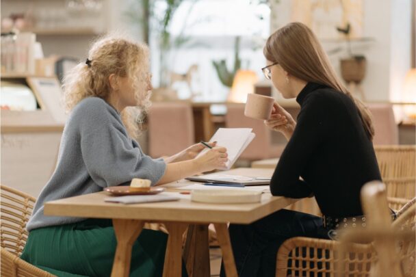 Twee vrouwen voeren een gesprek. Ze zitten aan tafel met koffie en wat papierwerk.