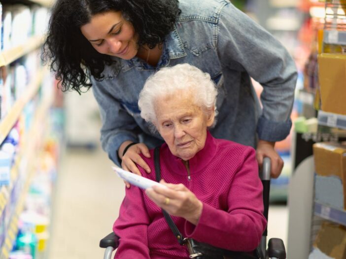 Oudere mevrouw zit in een rolstoel in de supermarkt. Jongere vrouw kijkt met haar mee op een stuk papier.