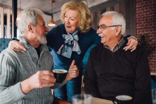 Twee oudere mannen drinken samen koffie en een oudere vrouw staat er gezellig bij.