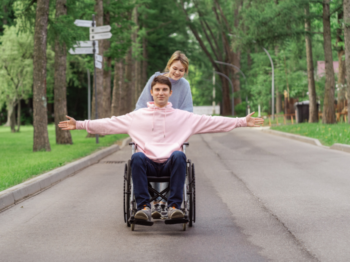 Jongen die in een rolstoel zit wordt geduwd door een meisje. Hij rijdt op de weg met armen gespreid onder de bomen.