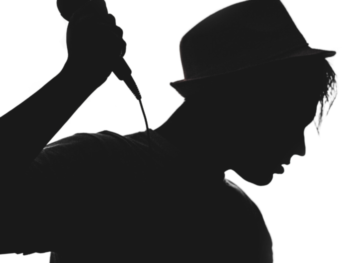 Zwart-wit foto met een man met een hoed. Hij is een pop artiest met een microfoon in zijn hand.