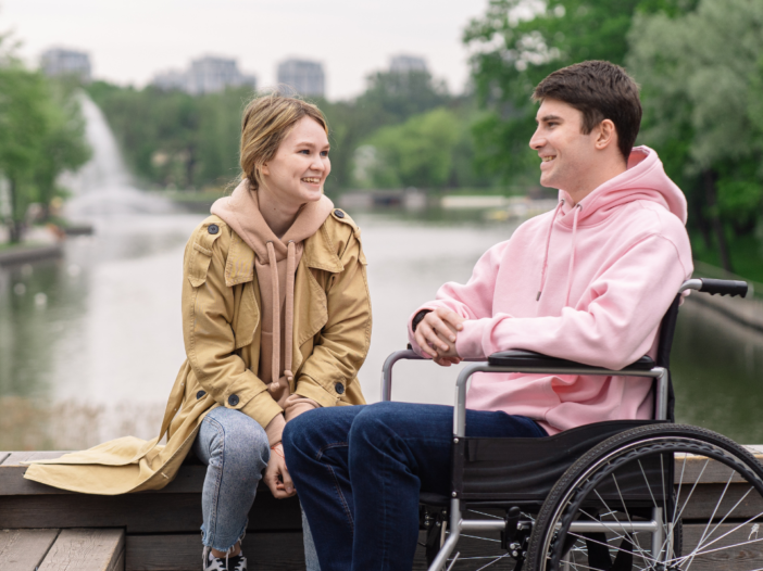 Jongen praat met meisje. Meisje met handen tussen haar benen en jongen in een rolstoel in een roze trui.