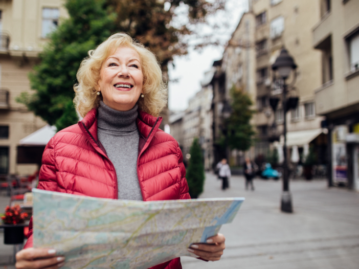 vrouw met rode bodywarmer loopt met een glimlach op haar gezicht door een stad. In haar handen heeft ze een kaart.