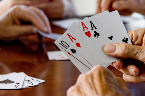 Er wordt een kaartspel gespeeld aan tafel. Je ziet een paar handen die kaarten vasthouden en een paar die een kaart van de stapel pakt. Er liggen ook kaarten op tafel