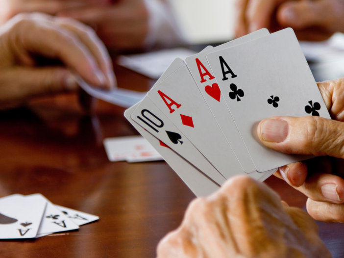Er wordt een kaartspel gespeeld aan tafel. Je ziet een paar handen die kaarten vasthouden en een paar die een kaart van de stapel pakt. Er liggen ook kaarten op tafel