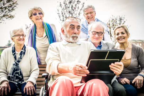Senioren zitten bij elkaar en kijken naar een Ipad. Ze kijken blij en verbaast.