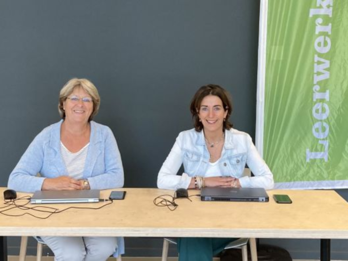 Twee vrouwen zitten naast elkaar aan een tafel. Er staat achter hun een doek met leerwerkloket erop en ze lachen.