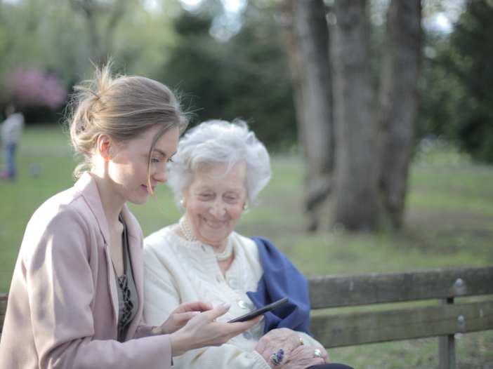 Oudere vrouw zit op een bankje buiten met jongere vrouw. Ze kijken naar een telefoon samen.