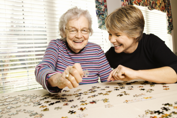 oudere vrouw zit aan tafel met jongere vrouw. Ze leggen een puzzel.