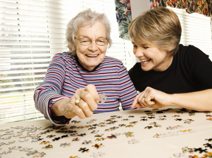 oudere vrouw zit aan tafel met jongere vrouw. Ze leggen een puzzel.