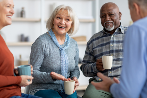 Vier senioren lachen samen en drinken een kop koffie. Ze zitten in een kring en de mevrouw in het midden is aan het lachen en aan het vertellen.