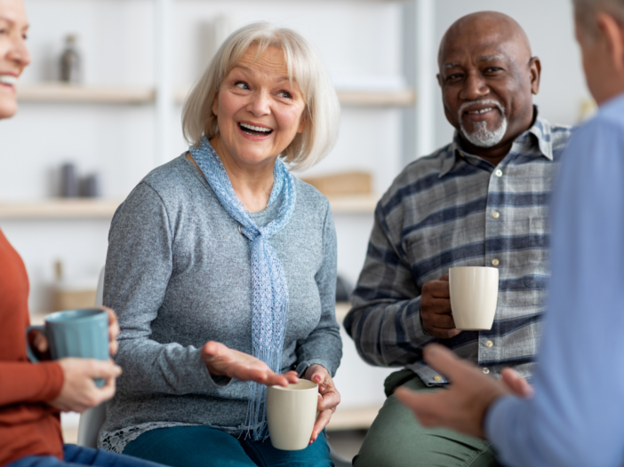 Vier senioren lachen samen en drinken een kop koffie. Ze zitten in een kring en de mevrouw in het midden is aan het lachen en aan het vertellen.