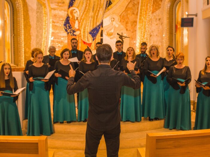 Een koor die in de kerk aan het zingen is. Ze hebben allemaal een groene rok aan en kijken naar de dirigent.