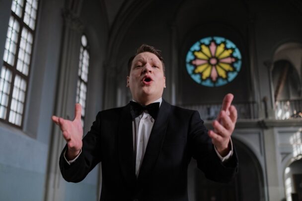 Een man zingt uit volle borst en drukt zichzelf ook uit met zijn handen. Hij staat in een kerk en heeft een zwart met wit pak aan.