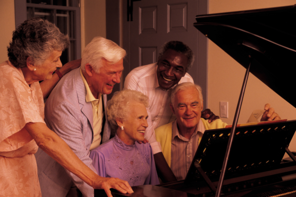 Groep oudere mensen die met z'n alle achter de piano staan. Witte mensen en donkere mensen achter de piano.