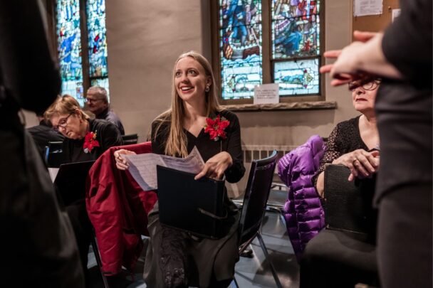 Lachende vrouw met notenboek in haar handen. Ze draagt een mooi zwart pak met een roos en ze zit in de kerk.