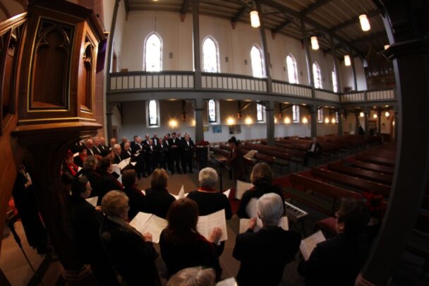 Ze zitten met z'n alle in een kerk te zingen. Ze hebben allemaal muziekbladen in hun handen.
