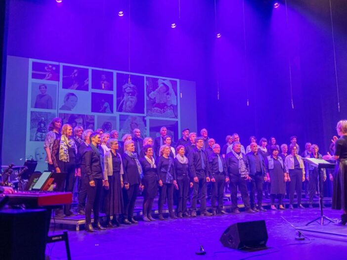 Impressie van het koor in Geldrop. Blauwe lichten en veel mensen op het podium die aan het zingen zijn.