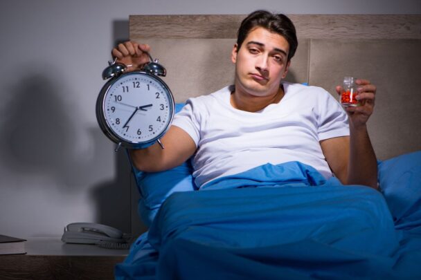 Jonge man ligt in zijn bed en kan niet slapen. Dit komt omdat hij slaapmiddelen in zijn hand heeft en dat werkt niet tegen een slaapprobleem.
