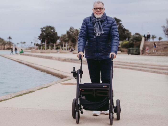 Een man staat buiten met een rollator. Hij heeft een jas en een sjaal aan.