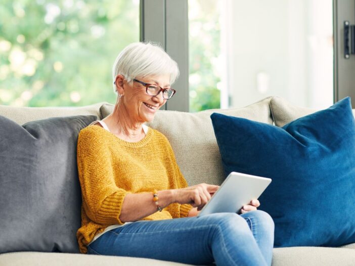 Mevrouw van oudere leeftijd zit op de bank. Ze klikt met haar vinger op het scherm van een ipad.