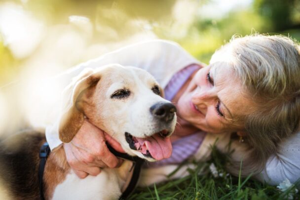Een vrouw van oudere leeftijd ligt op het gras. Ze knuffelt met een hond, zijn tong hangt uit zijn bek.