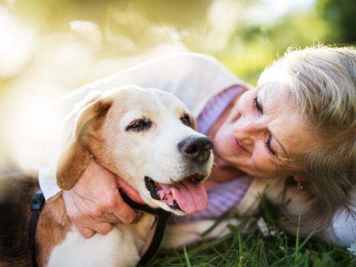 Een vrouw van oudere leeftijd ligt op het gras. Ze knuffelt met een hond, zijn tong hangt uit zijn bek.