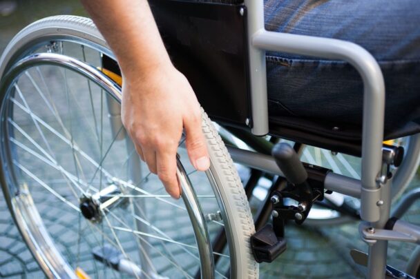 Er zit iemand in een rolstoel. Je ziet een zij-aanzicht met het wiel, een stuk van de stoel en een hand.