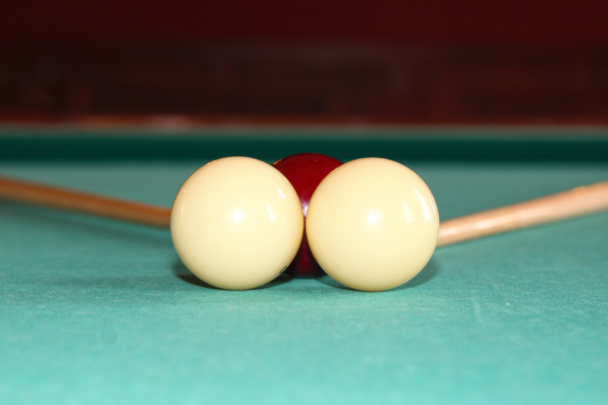 Twee biljartballen worden allebei geslagen met een biljartstok. De ballen liggen op een groene biljartmat en zijn wit.