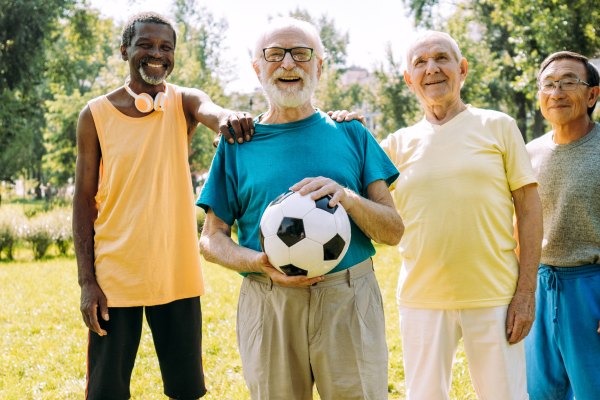 vier mannen van oudere leeftijd staan buiten op een grasveld. Ze hebben een bal vast en lachen.