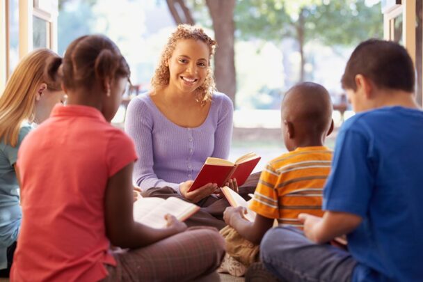Vrouw leest voor aan kinderen. Ze zitten gezellig in een kring en de kinderen lezen aandachtig mee.