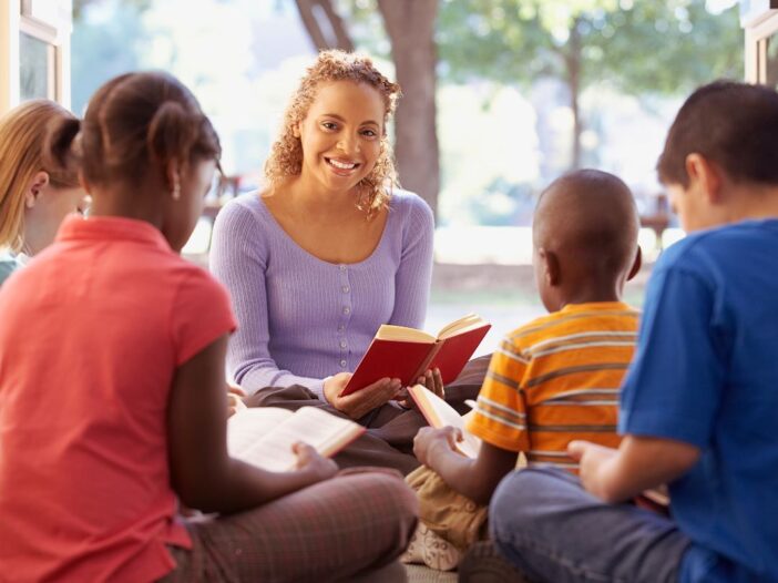 Vrouw leest voor aan kinderen. Ze zitten gezellig in een kring en de kinderen lezen aandachtig mee.