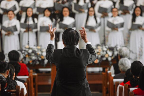 Dirigent met zijn handen de lucht in. Voor hem staat een koor met allemaal witte kleren aan.