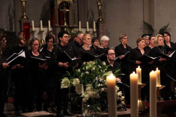 Zanggroep in Hoogeloon zingen allemaal uit een boekje. Ze hebben allemaal zwarte kleren aan en staan in de kerk. De kaarsen staan ook aan.