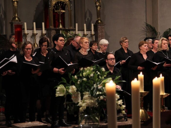 Zanggroep in Hoogeloon zingen allemaal uit een boekje. Ze hebben allemaal zwarte kleren aan en staan in de kerk. De kaarsen staan ook aan.