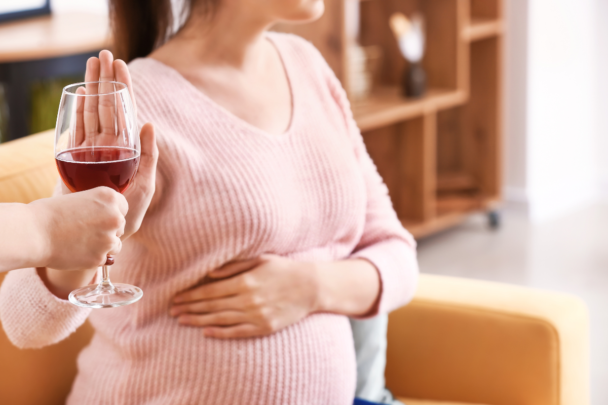 Een zwangere vrouw met een roze shirt aan zegt nee tegen een glas rode wijn. Ze zegt nee omdat ze zwanger is en zwangere vrouwen mogen geen alcohol drinken.