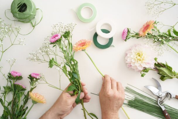 Twee handen pakken losse bloemen bij elkaar. Op tafel liggen meer bloemen en lint.