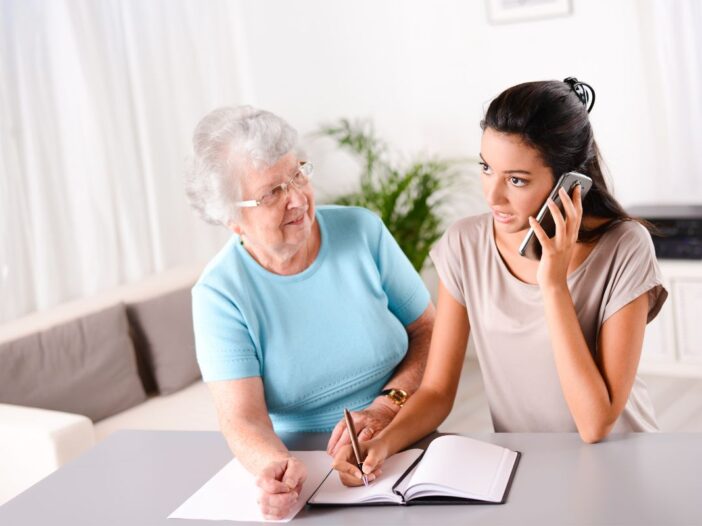 Jongere vrouw is aan het bellen en in een schrift aan het schrijven. Een oudere vrouw kijkt naar haar.