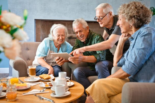 Vier ouderen maken samen gebruik van een tablet en lachen. Op tafel staan glazen drinken.