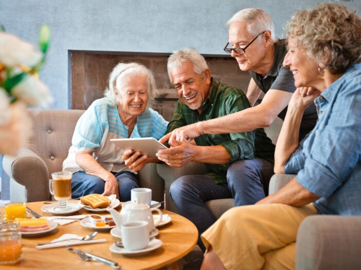 Vier ouderen maken samen gebruik van een tablet en lachen. Op tafel staan glazen drinken.