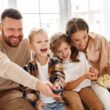 Man en vrouw zitten samen met twee kinderen op de bank. Ze hebben plezier en eten popcorn.