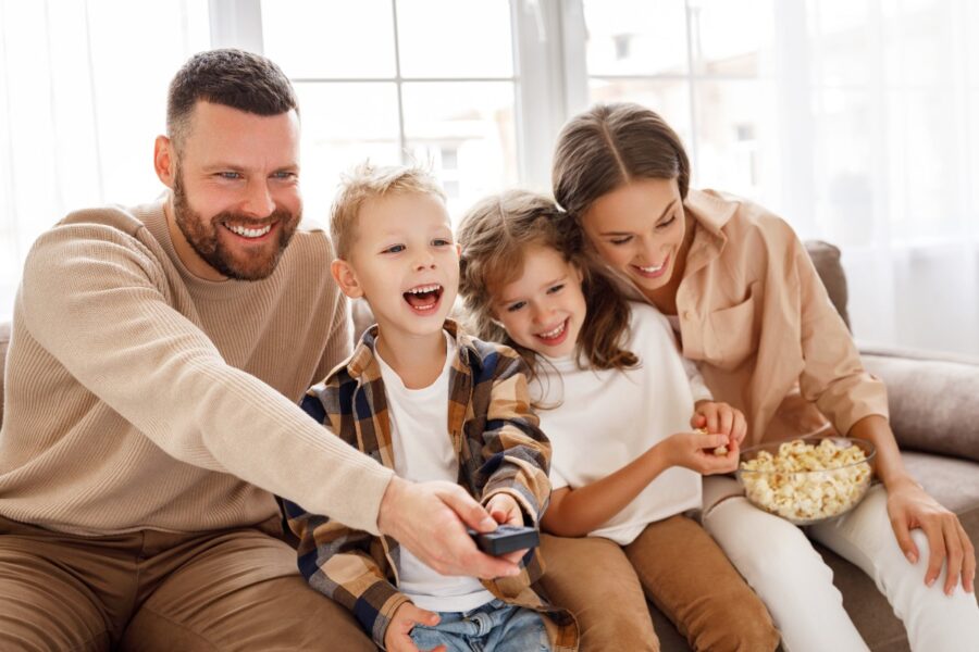 Man en vrouw zitten samen met twee kinderen op de bank. Ze hebben plezier en eten popcorn.