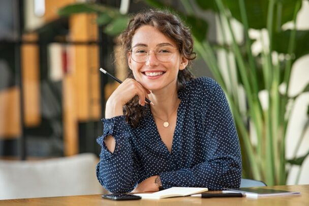 Jonge vrouw met bril en donkerblauwe blouse kijkt lachend in de camera.