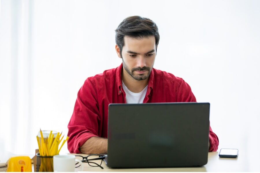 Man met rode blouse aan maakt gebruik van een laptop. Hij zit aan een tafel.