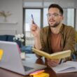 Man met bril zit achter laptop met een boek en pen in zijn handen.