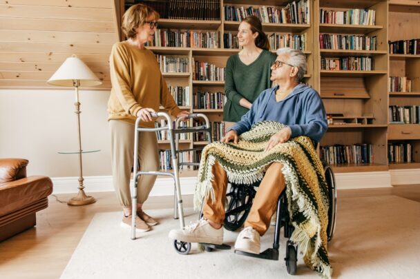 Twee ouderen krijgen hulp in huis van een mantelzorger. De oude man zit in een stoel en de oudere vrouw staat er naast met een looprek.