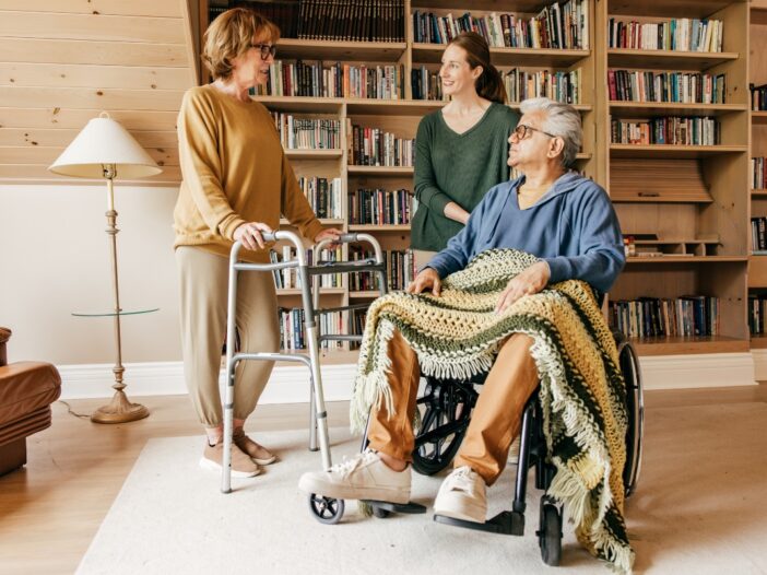 Twee ouderen krijgen hulp in huis van een mantelzorger. De oude man zit in een stoel en de oudere vrouw staat er naast met een looprek.