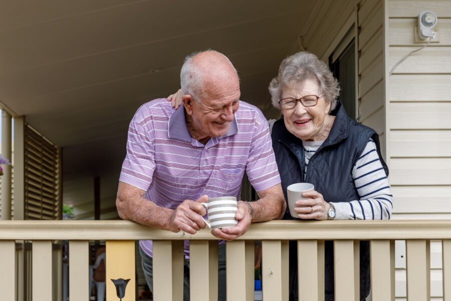 Oudere man en vrouw staan samen op een veranda en genieten van een kop koffie.