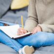 Vrouw zit op de bank in spijkerbroek met een schrift op schoot. Ze is aan het schrijven.